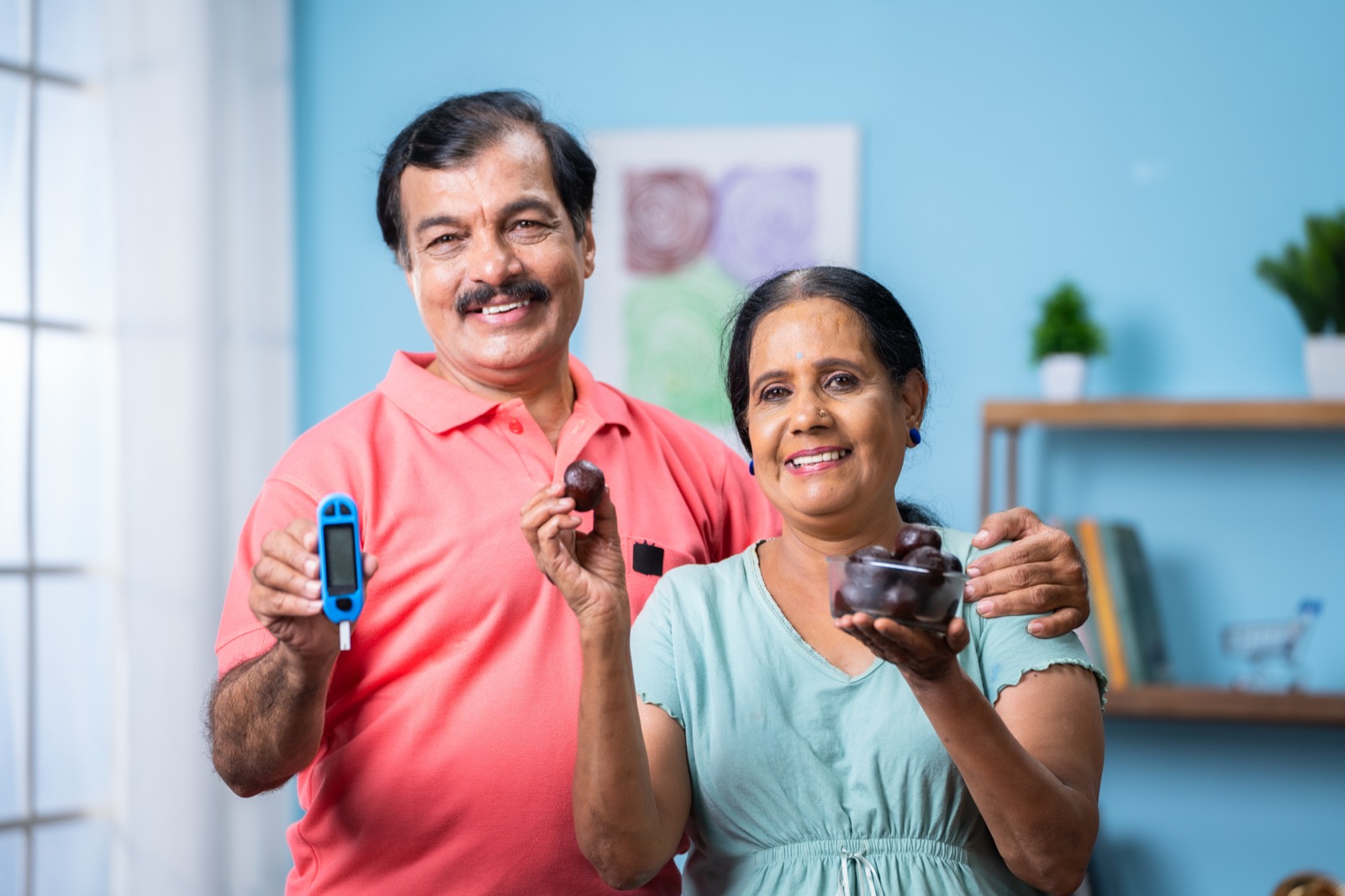 A smiling Indian senior couple at home holding a glucometer